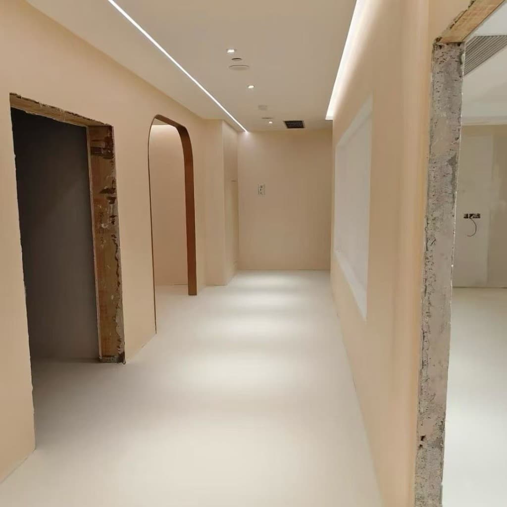 Hotel corridor with arched openings, peach-cream limewash walls and a continuous warm-white microcement floor, recessed LED light strip in the soffit