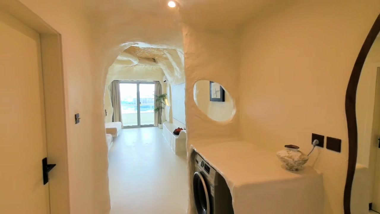 Guest suite entry framed as a sculpted cave arch with an oval mirror cut directly out of the microcement wall, washing-machine niche on the right and city-view balcony at the end
